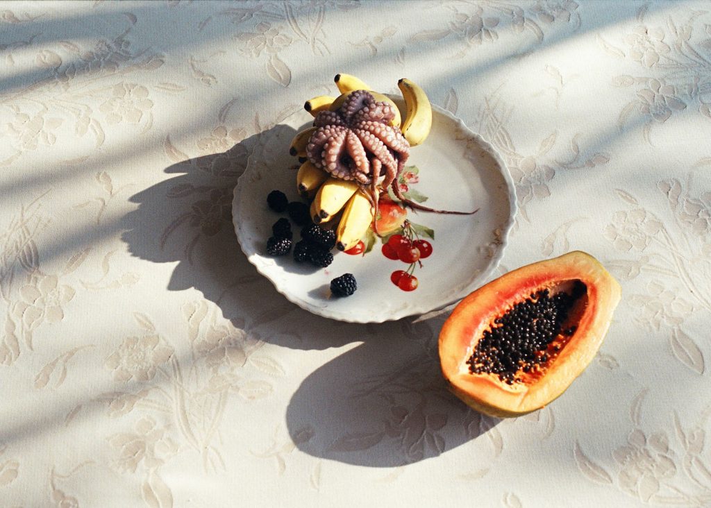 Overhead shot of a plate with bananas, blackberries, and an octopus, alongside a halved papaya on a floral tablecloth.