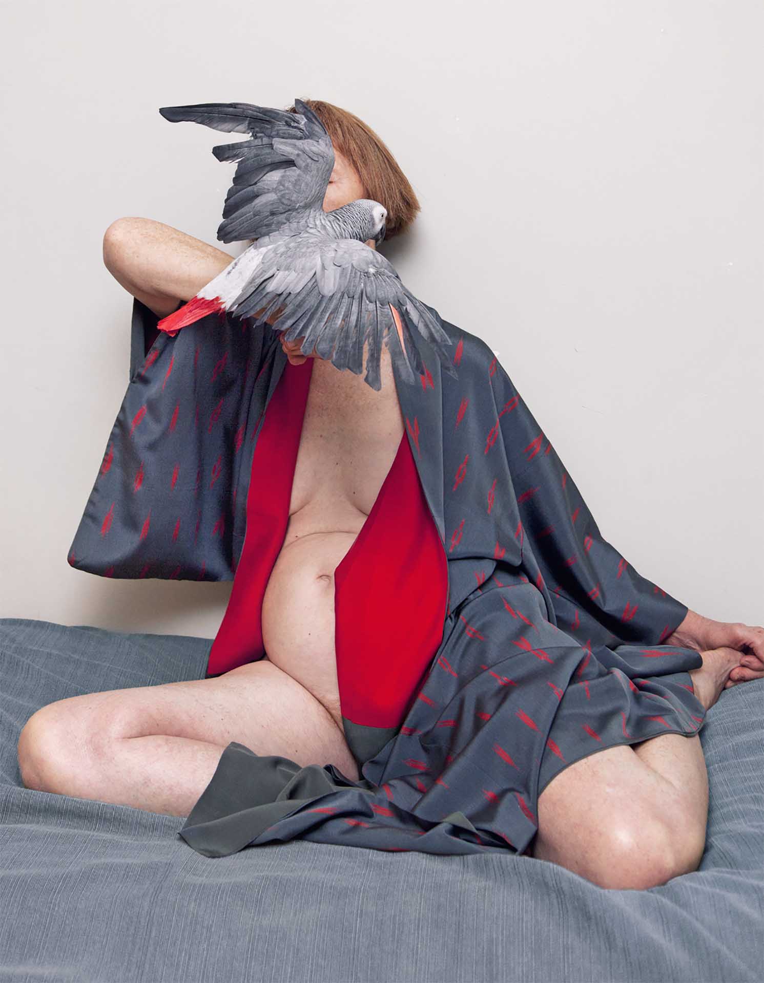 A woman in a robe sits cross-legged with a gray parrot perched on her shoulder in a brightly lit indoor setting on a blue-gray bedspread.
