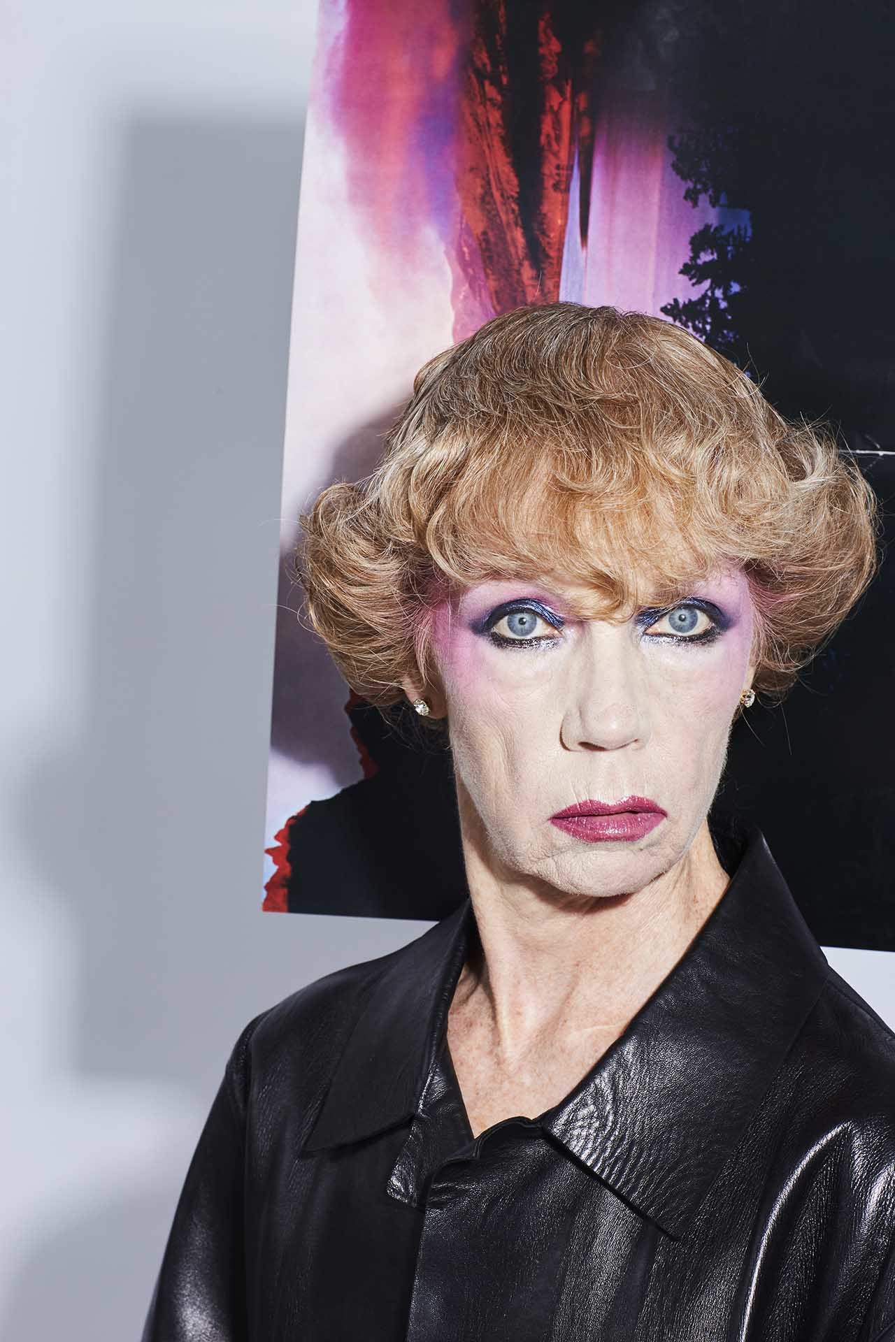 Portrait of an older woman with pink eyeshadow and short blonde hair wearing a black leather jacket against a vibrant backdrop.
