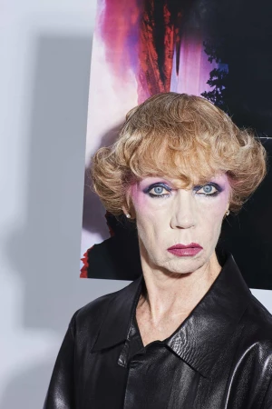 Portrait of an older woman with pink eyeshadow and short blonde hair wearing a black leather jacket against a vibrant backdrop.