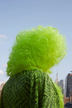 Person with bright lime green curly wig wearing textured green patterned top against clear blue sky