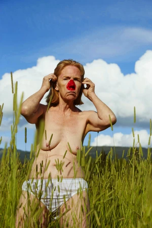 Elderly woman with a red clown nose stands in a grassy field under a bright blue sky with fluffy white clouds on sunny day.