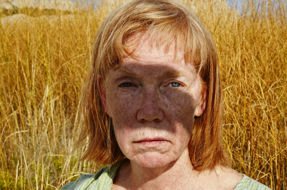 Close-up portrait of a mature woman with red hair and freckles, gazing directly at the viewer, standing in a field of golden grass.