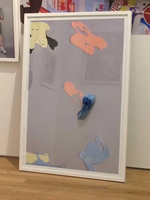Framed artwork featuring abstract shapes and colors on a gray background, exhibited indoors, creating a visually engaging display.