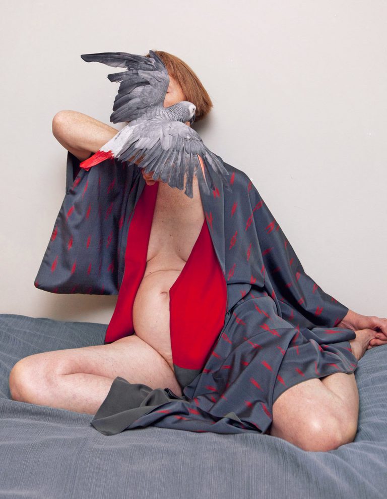 Adult female in kimono on bed with African grey parrot perched on shoulder, striking a unique pose in an intimate, indoor setting.