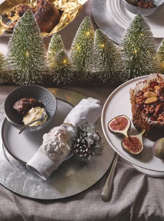 Festive table setting featuring a small Christmas tree forest, dessert in a bowl, a napkin ring, and a fig cake on plates.