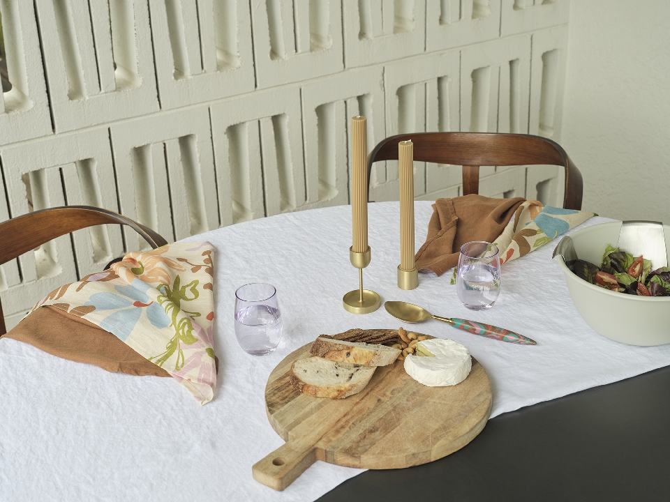Table setting featuring candles, water glasses, bread, cheese, and salad, arranged on a white tablecloth with a modern, minimalist backdrop.