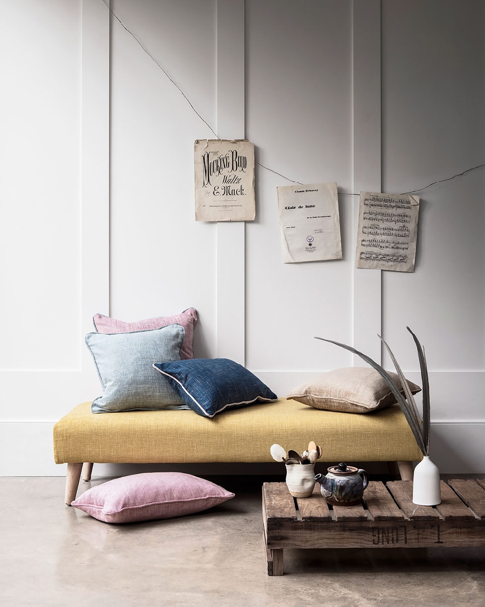 Interior scene with a yellow upholstered bench topped with linen cushions, vintage sheet music hanging on the white paneled wall.