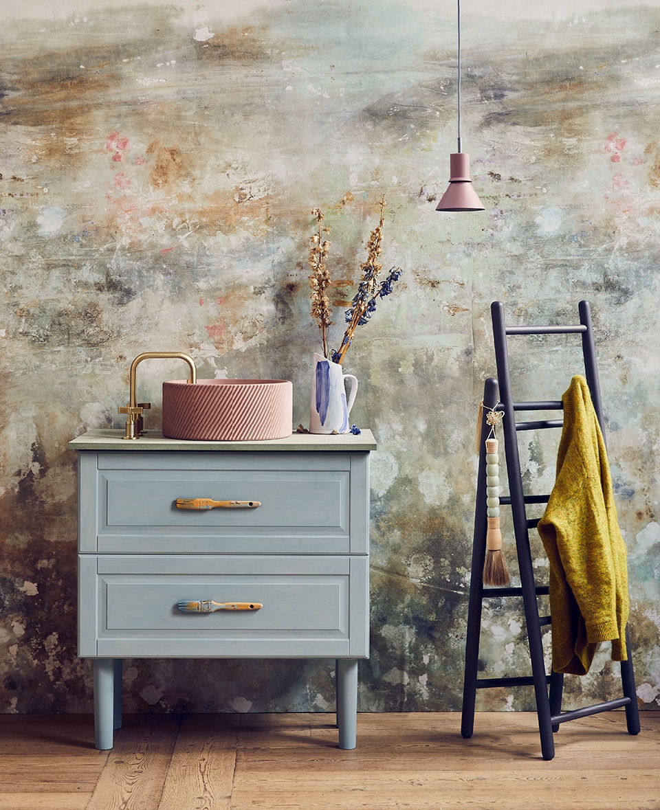 Bathroom interior featuring a vanity with a pink sink, gold faucet, dried flowers in a vase, and a ladder with a yellow towel.