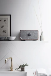 A rectangular speaker with fabric grille and wooden frame rests on a white floating shelf styled by Lucy Gough.