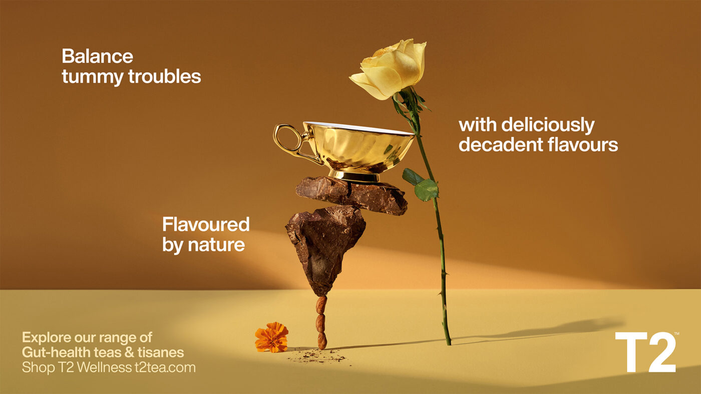 Still life food and beverage product shot featuring a yellow rose, gold teacup, chocolate, almonds, and orange flower on a beige background.