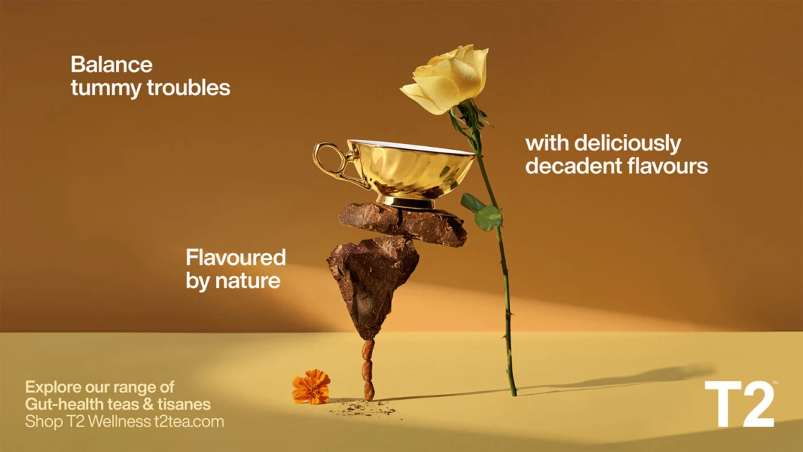 Still life food and beverage product shot featuring a yellow rose, gold teacup, chocolate, almonds, and orange flower on a beige background.