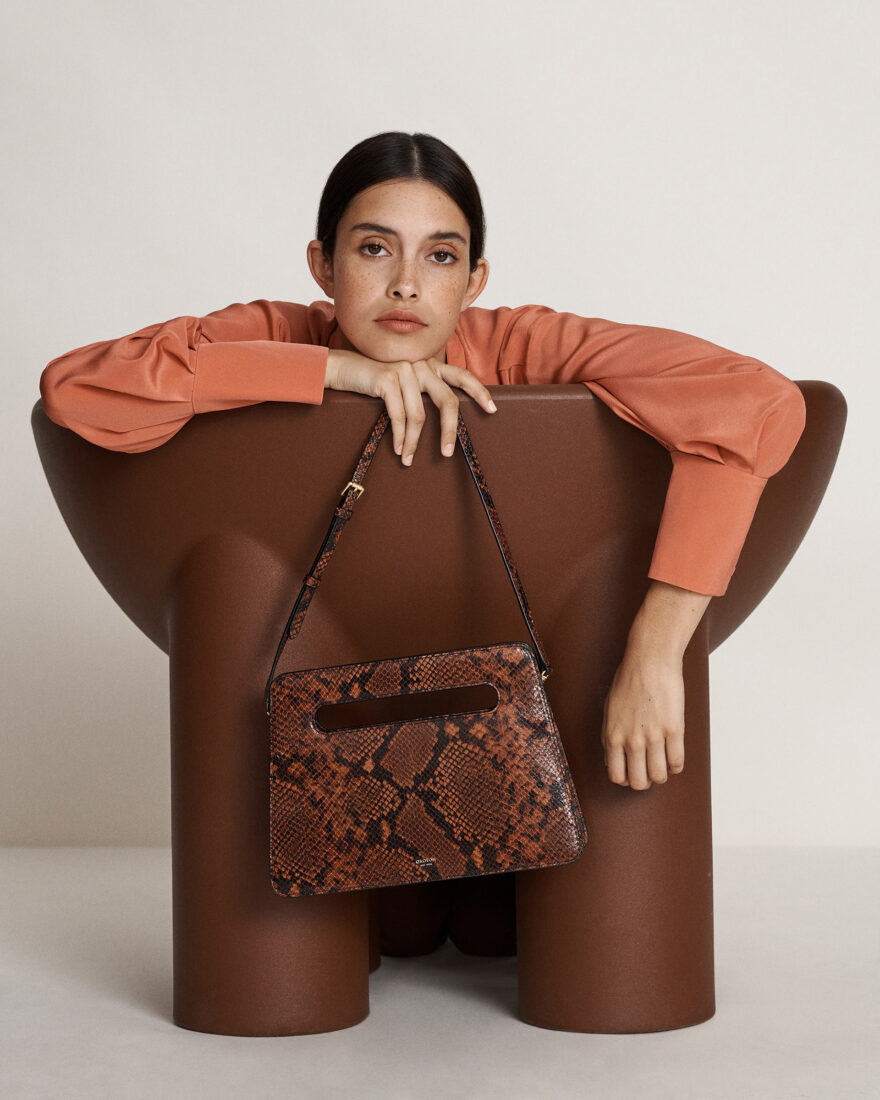 Studio shot of a woman in a peach sweater with a snakeskin handbag, seated in a brown chair against an off-white backdrop.