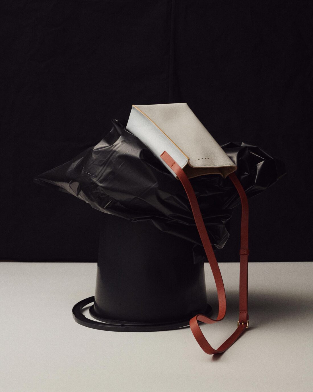 Studio shot of a white Marni handbag with a brown shoulder strap, posed on top of a black trash can against a black and white backdrop.