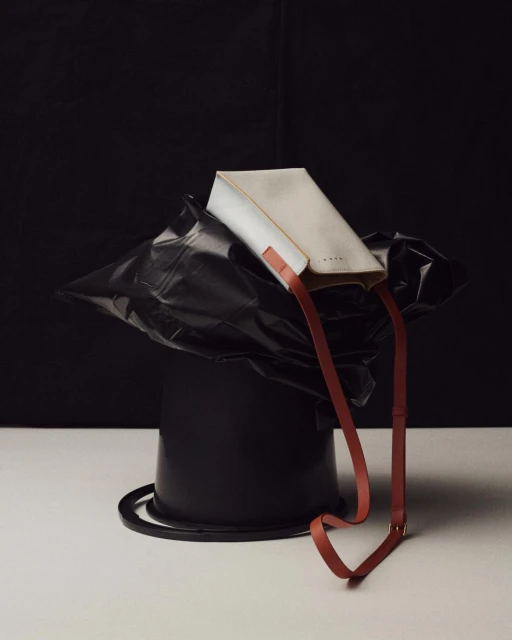 Studio shot of a white Marni handbag with a brown shoulder strap, posed on top of a black trash can against a black and white backdrop.