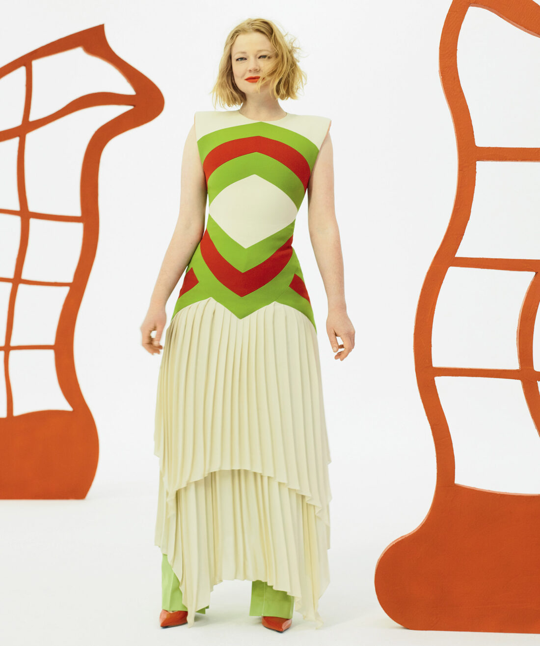 Woman in a retro-style color-blocked dress posing in a bright studio with painted orange cut-out shapes for a fashion editorial.