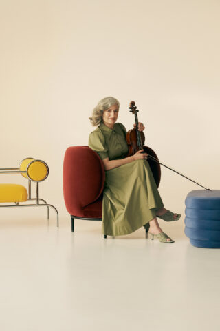 Mature woman seated holding violin in photo studio setting, wearing green dress and surrounded by modern furniture pieces.