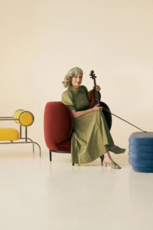 Mature woman seated holding violin in photo studio setting, wearing green dress and surrounded by modern furniture pieces.