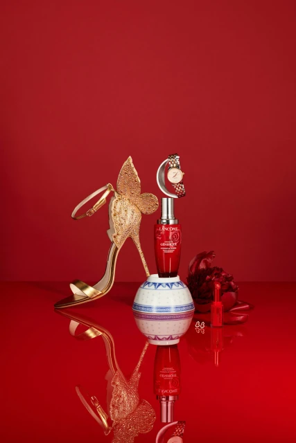 Still life featuring a Lancôme Génifique serum bottle, golden high heel, and a watch on a reflective surface against a red backdrop.