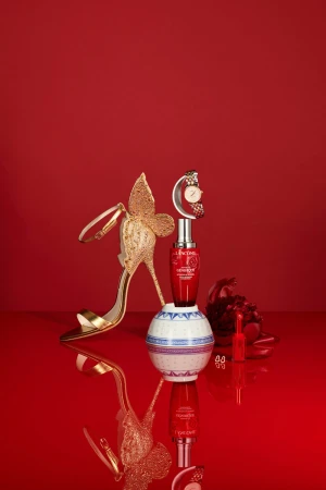 Still life featuring a Lancôme Génifique serum bottle, golden high heel, and a watch on a reflective surface against a red backdrop.