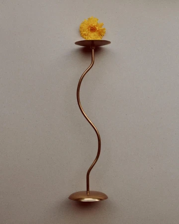 Minimalist studio shot of a single yellow gerbera daisy displayed on a tall, curving copper candlestick against a light gray background.