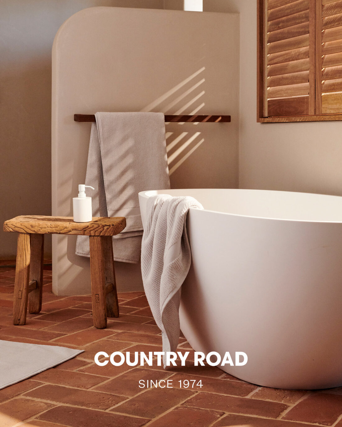 Bathtub in a neutral bathroom setting with a wooden stool, a towel rack holding a towel, and natural light coming through window shutters.