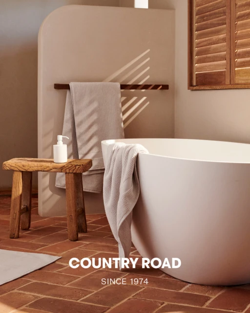 Bathtub in a neutral bathroom setting with a wooden stool, a towel rack holding a towel, and natural light coming through window shutters.