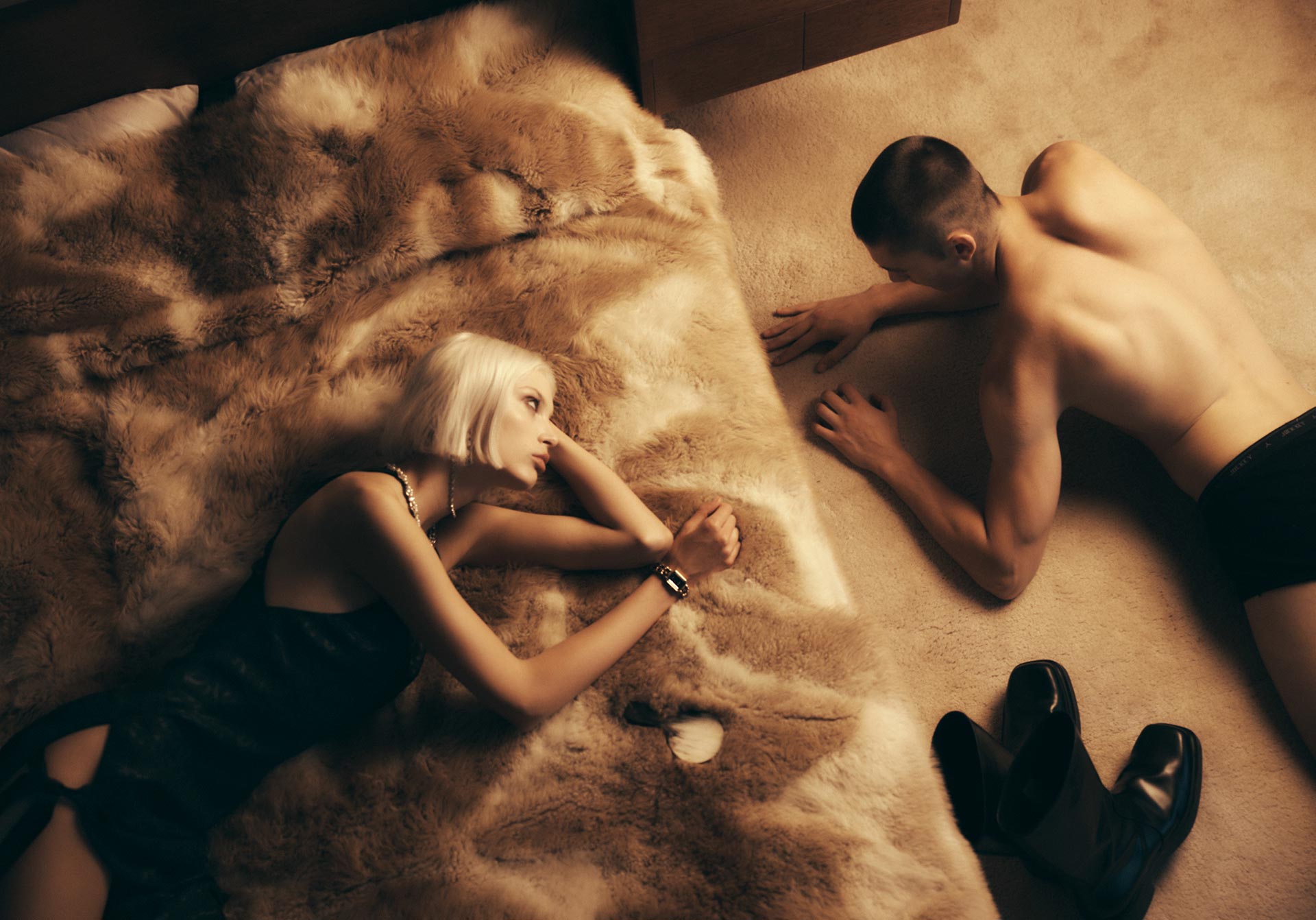 Blonde woman in dress and man in underwear lying on floor with shoes. Couple indoors on carpet, with furry throw on bed.