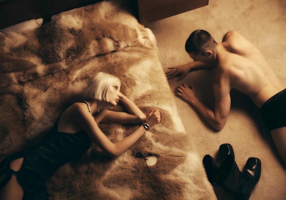 Blonde woman in dress and man in underwear lying on floor with shoes. Couple indoors on carpet, with furry throw on bed.