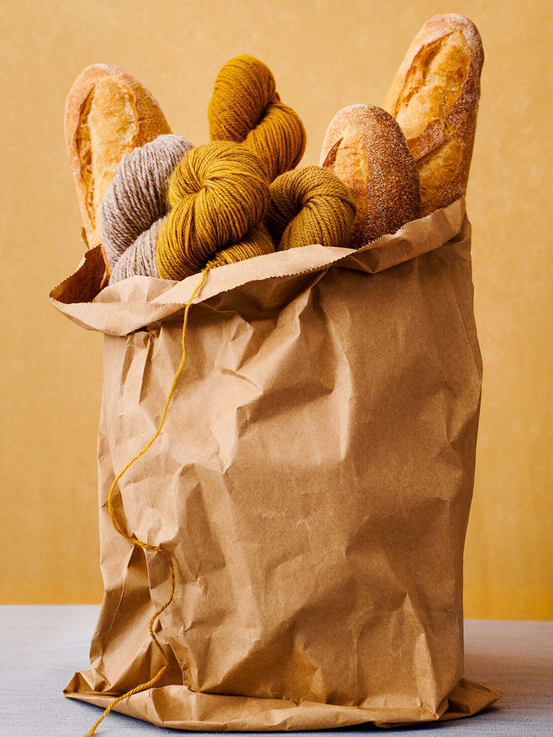 Two crusty baguettes and balls of golden and gray yarn are arranged inside of a brown paper grocery bag on a light gray surface.