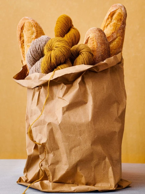Two crusty baguettes and balls of golden and gray yarn are arranged inside of a brown paper grocery bag on a light gray surface.