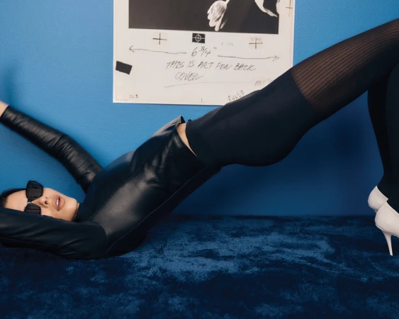 Model in black leather outfit and sunglasses bridging over blue shag carpet with framed print behind.