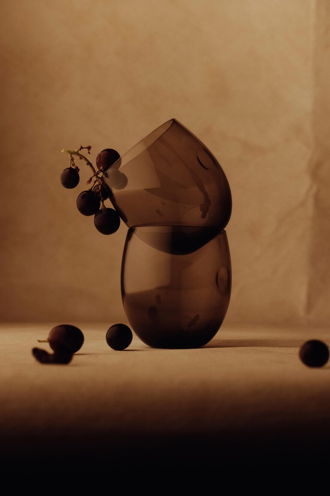 Still life studio photograph of a stack of two rounded smoky glass vases, accompanied by a sprig of dark grapes against a neutral background.