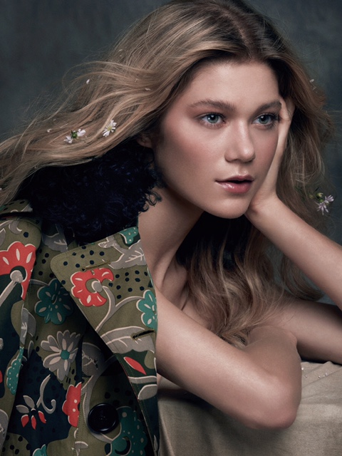 Portrait of a blond woman with flowers in her hair wearing an olive green floral coat in a studio setting with a grey backdrop.