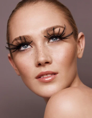 Close up portrait of a young woman with dramatic long false eyelashes and light freckles against a neutral background.