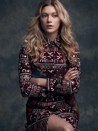 Portrait of a blonde woman with arms crossed wearing a patterned burgundy dress against a textured dark grey backdrop.