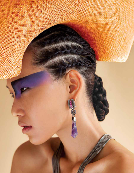 Close-up of a stylish woman with unique makeup and braided hair, wearing a large orange hat and statement earrings in a studio setting.