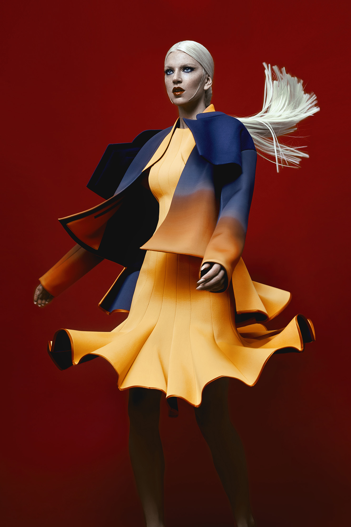 Fashion model poses in a futuristic yellow-and-blue dress with blonde hair against a vibrant red background in a studio setting.