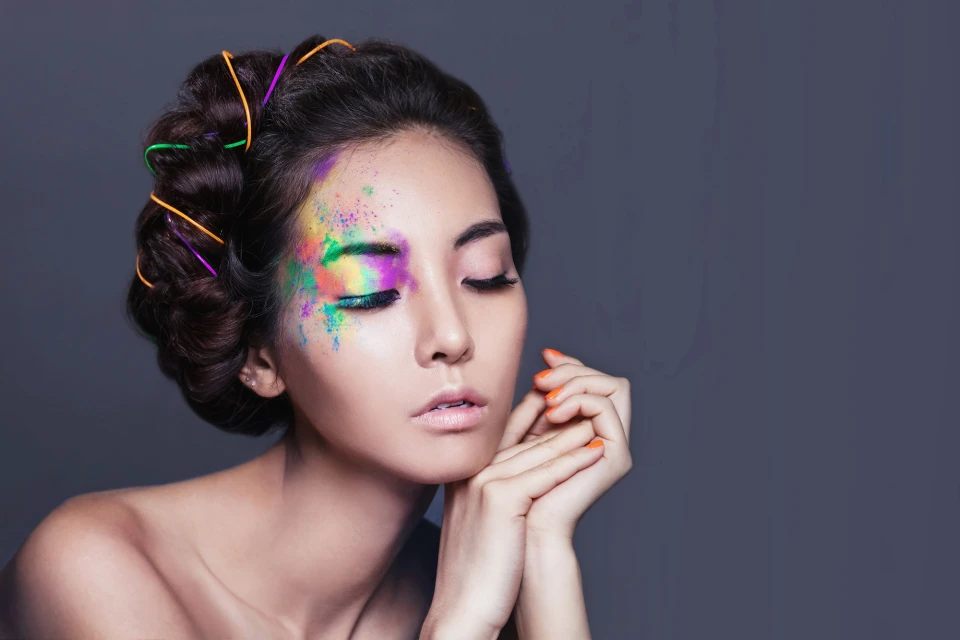 Close-up portrait of a woman with colorful face paint, soft lighting, closed eyes and hair accessories on a dark gray background.