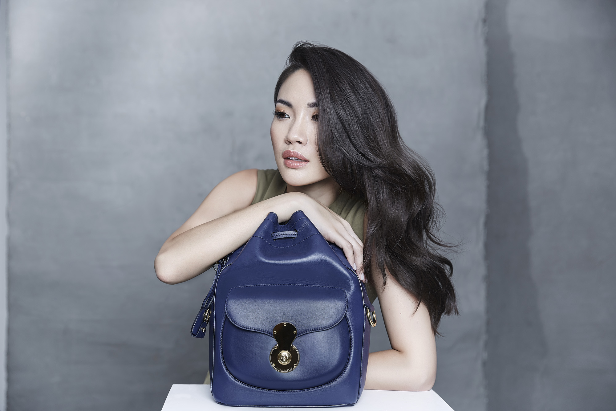 Attractive adult woman poses with a stylish navy-blue leather bucket bag in a fashion studio against a textured grey backdrop.