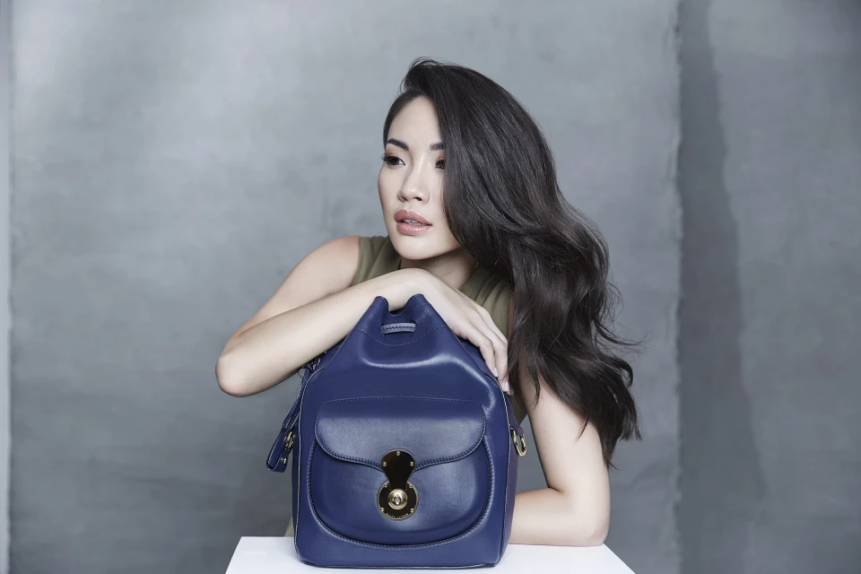 Attractive adult woman poses with a stylish navy-blue leather bucket bag in a fashion studio against a textured grey backdrop.