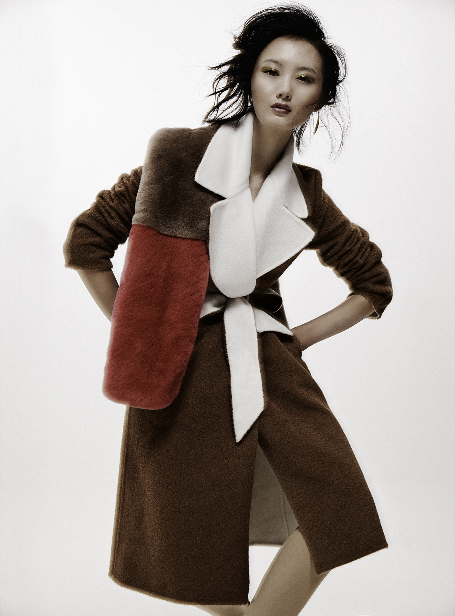 Asian woman poses in a brown coat with red and brown panels, cinched with a white belt, against a clean white backdrop in a fashion shot.