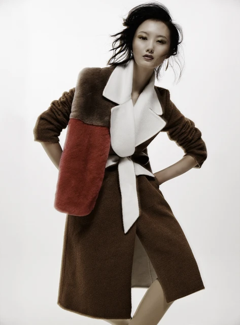Asian woman poses in a brown coat with red and brown panels, cinched with a white belt, against a clean white backdrop in a fashion shot.
