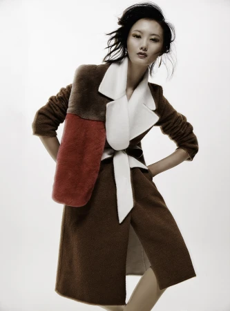 Asian woman poses in a brown coat with red and brown panels, cinched with a white belt, against a clean white backdrop in a fashion shot.