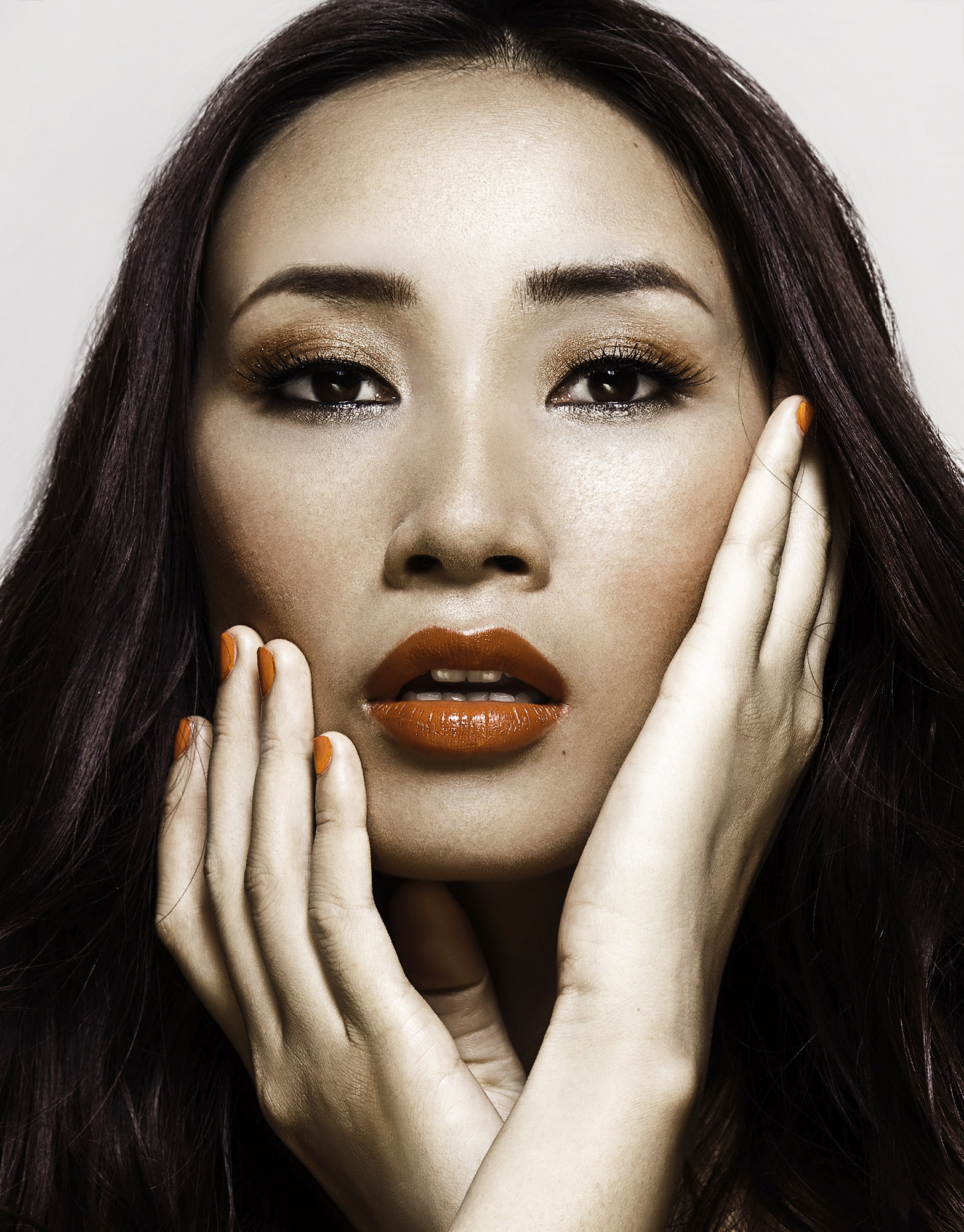 Close-up portrait of an Asian woman with vibrant orange lipstick and matching nail polish, posing with her hands framing her face.