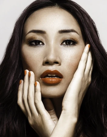 Close-up beauty portrait of an East Asian woman with dark hair, orange lipstick, and vibrant orange nails framing her face.