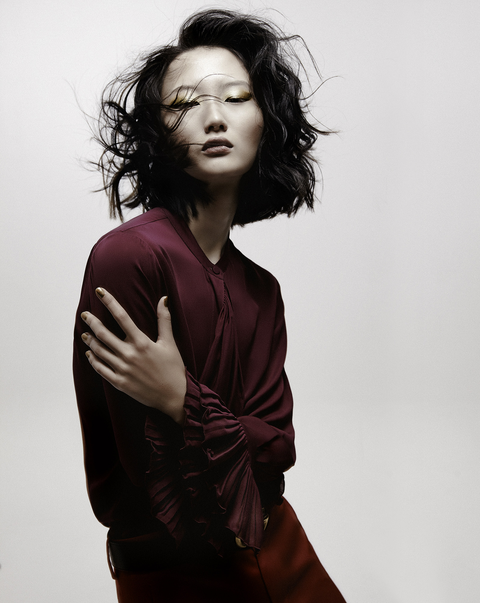 Asian woman with gold eyeshadow poses in a photo studio. She wears a red-purple top, red skirt, and has short black hair.