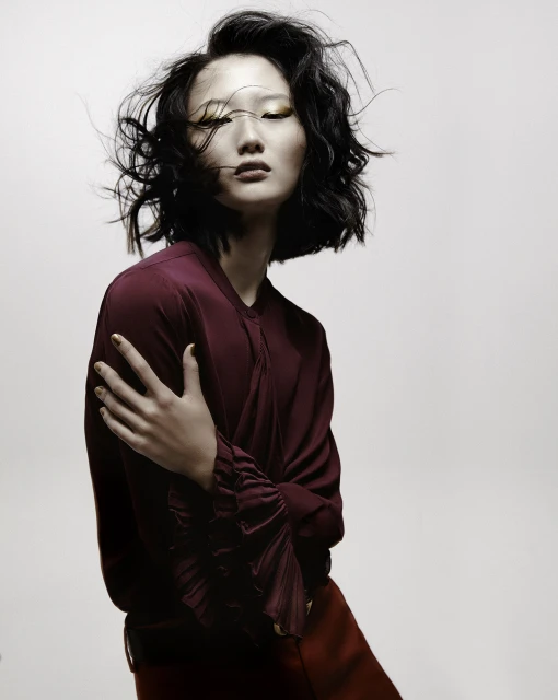 Asian woman with gold eyeshadow poses in a photo studio. She wears a red-purple top, red skirt, and has short black hair.