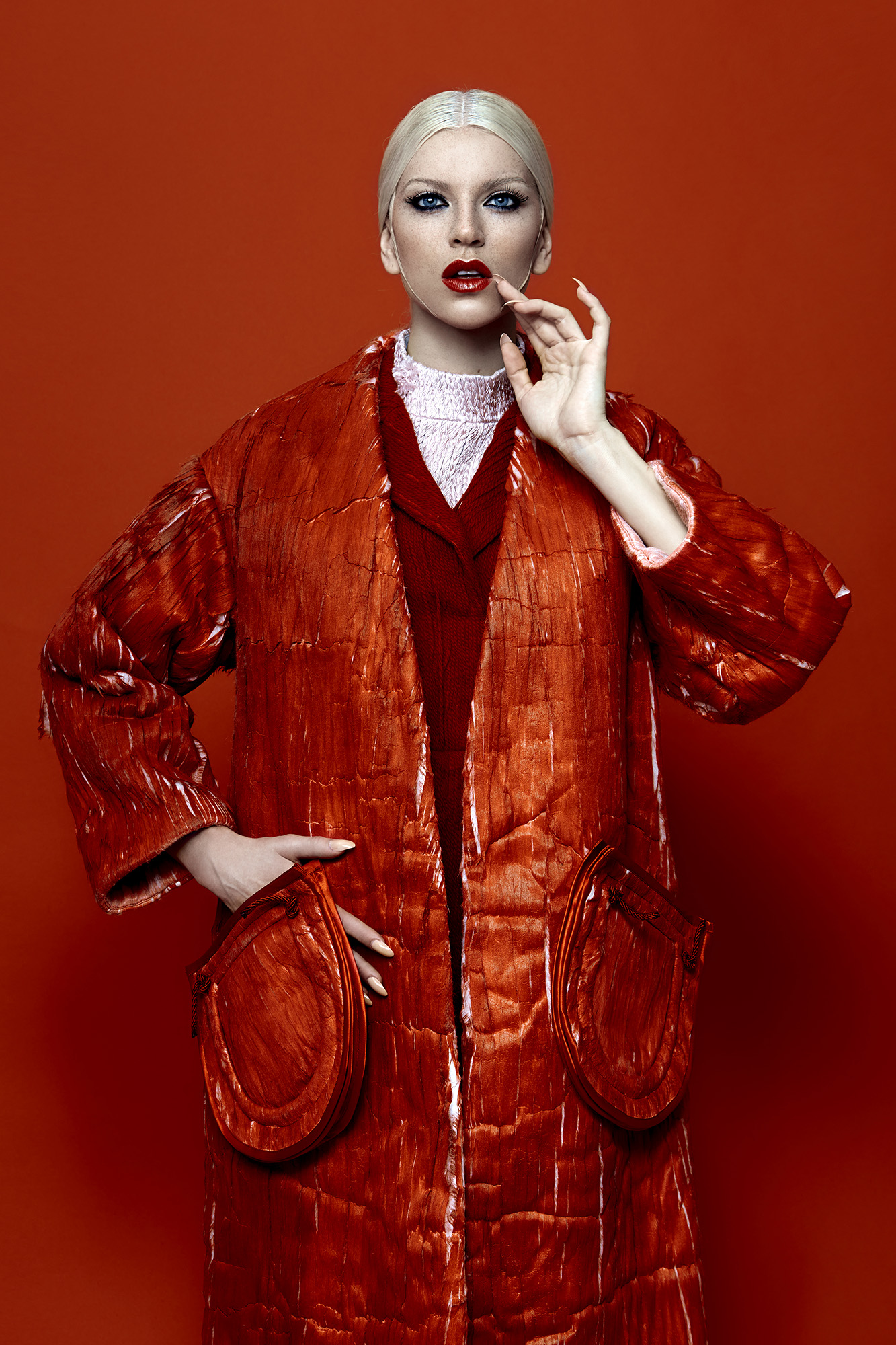 Fashionable woman with pale blonde hair, wearing a red overcoat with puffy pockets against a solid red background in a studio shot.