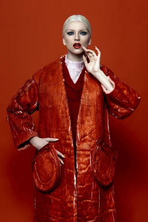 Fashionable woman with pale blonde hair, wearing a red overcoat with puffy pockets against a solid red background in a studio shot.